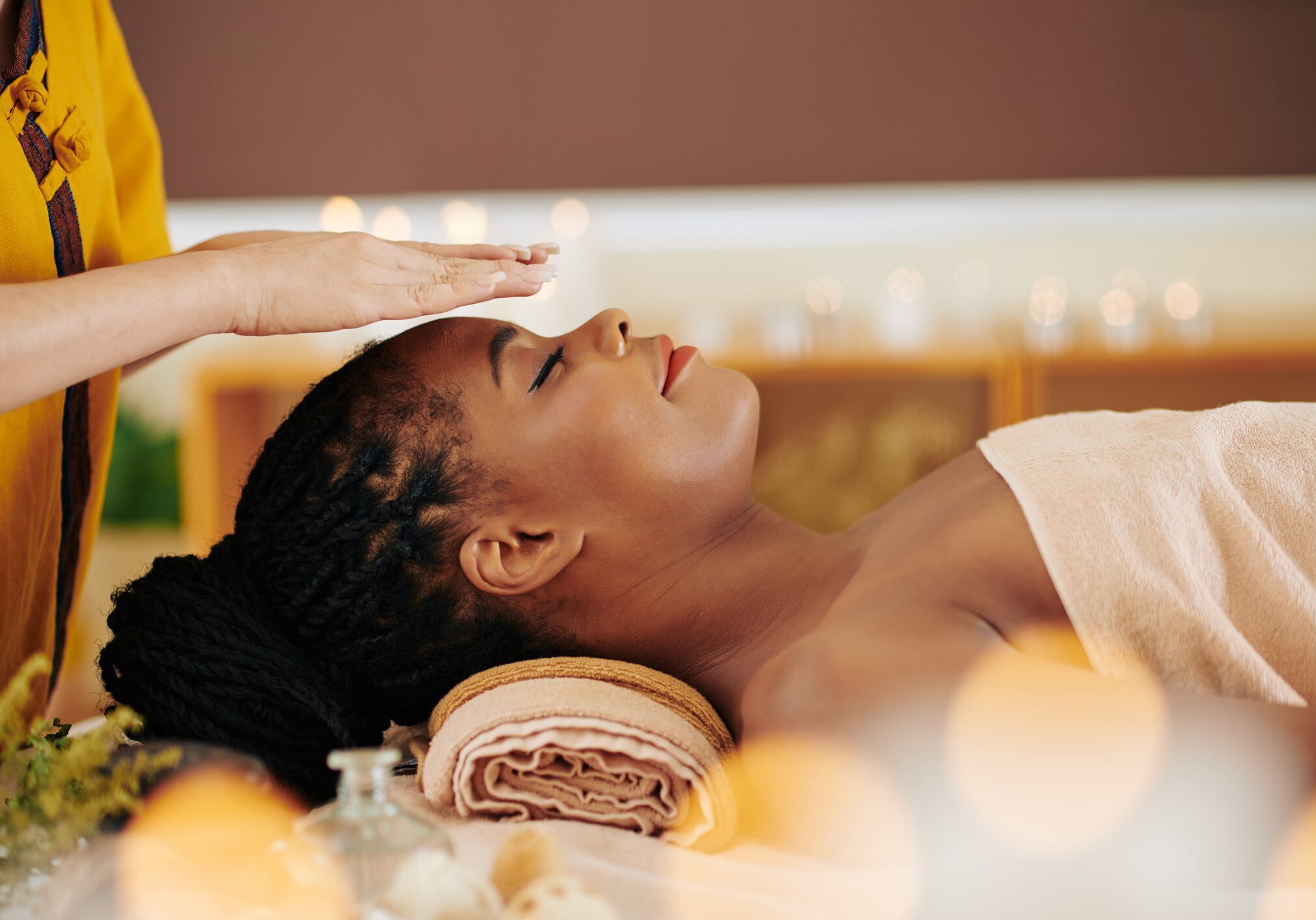 Therapist holding hands over head of pretty young Black woman to transfer energy during healing reiki session